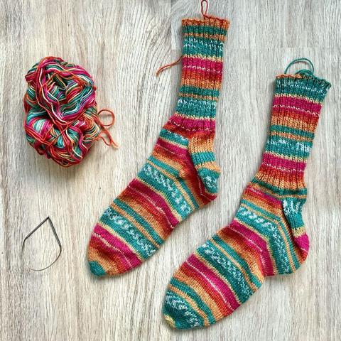 Flat lay view of 2 socks in self-striping yarn in yellow, orange, pink, and teal, on a light wooden background. The tiny circular needle, and the remaining yarn are also visible.