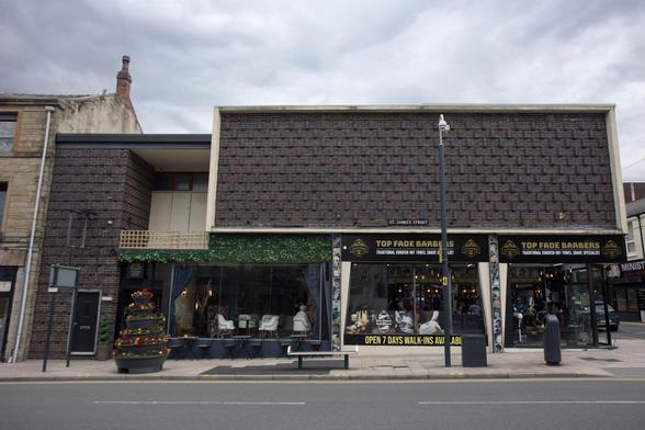 A modern barbershop with large glass windows and TOP FADE BARBERS signs above. Outdoor seating with tables and chairs is in front, along with a small decorative tree adorned with orange ornaments. The building has a brick façade and faces a quiet street.