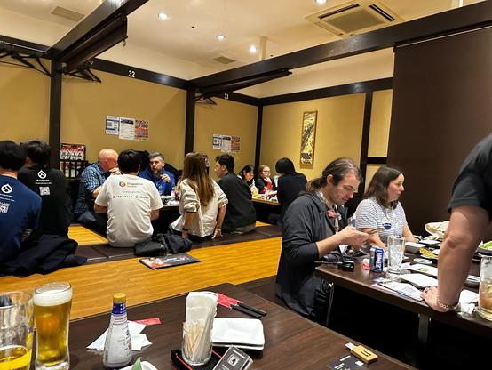A busy izakaya filled with DrupalCon participants sitting on tatami-style platforms and benches, talking and eating in small groups. Beer glasses, shared dishes, and casual conversations fill the room, giving a sense of community, warmth, and the relaxed energy of a conference evening in Japan.