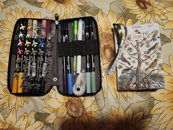 A picture of the planner whose cover has a tree with a different bird in every branch. It has a sage green ribbon and a white ribbon with green leaves. Next to the planner is a cloth marker organizer full of double sided markers, with holographic plane stickers, and a whiteout tape dispenser.