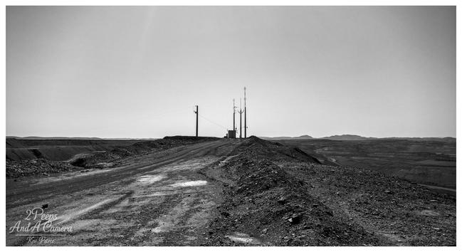 A low angle, black and white photograph signed by Kev Peirce. A rough, dirt track littered with rubble leads up a steep embankment toward a cluster of utility poles and a radio communication tower in the distance.

The foreground is heavily textured with shadows and loose rock. The horizon is wide and flat, with low hills visible in the far background under a bright, clear sky.

The landscape suggests an arid, remote, or mining affected area, likely near Leigh Creek.