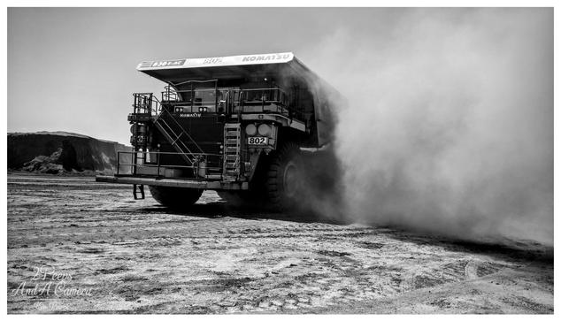 A dynamic, low angle, black and white photograph signed by Kev Peirce. A massive Komatsu 830E-AC haul truck, identified by its model number and '802' on the side, is seen from the rear quarter.
The truck is driving on a dusty, cracked dirt surface, generating a huge, thick cloud of dust that billows dramatically to the right side of the frame.
In the background, there is a large, sheer embankment of excavated material. The image captures the scale and activity of a major mining operation.