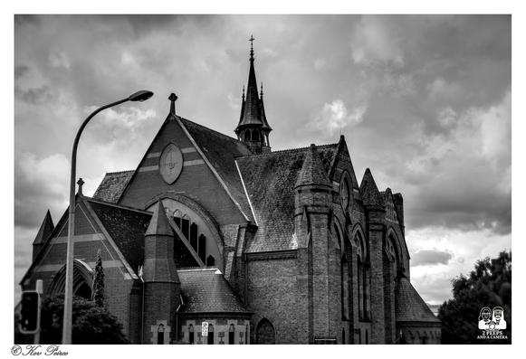 A striking black and white, low angle photograph of a historic church in Launceston, Tasmania.

The building is constructed of dark brick in a Gothic Revival style, featuring steep slate roofs, buttresses, and tall, narrow arched windows.

A small spire topped with a cross rises from the center of the roofline. The dramatic sky is filled with heavy, textured clouds, creating a strong contrast with the architecture.

A modern streetlamp and some dark foliage are visible on the left side, slightly framing the building.