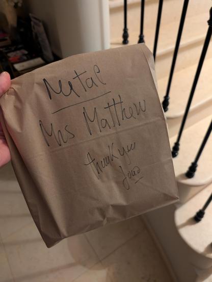 A brown bag with black permanent marker writing. On the bag reads "Metal" (room name), "Mrs. Matthew, thank you, João" 