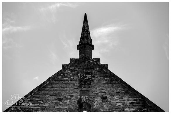The image focuses tightly on the brick gable end and central stone spire of a historic ruined building, likely the church or main structure at Port Arthur, Tasmania.

The rough, textured brickwork fills the base of the frame, converging into a central stone spire that points dramatically upwards into a bright, cloudy sky.