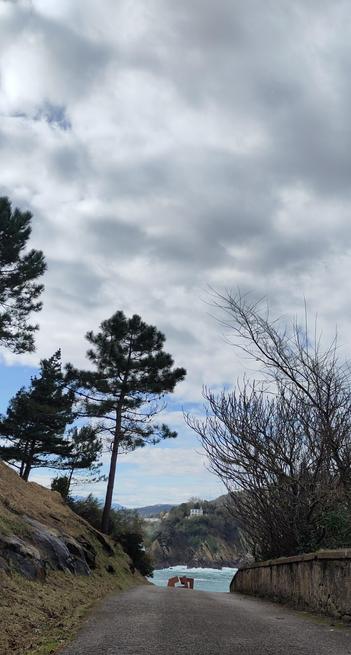 Der Blick führt über einen asphaltierten Weg, von Bäumen gesäumt, an einem Stück einer Skulptur vorbei aufs Meer. Felsen einer Insel sind dort zu sehen, obenauf ein weißes Gebäude. Der Himmel darüber leicht blau aber bewölkt.