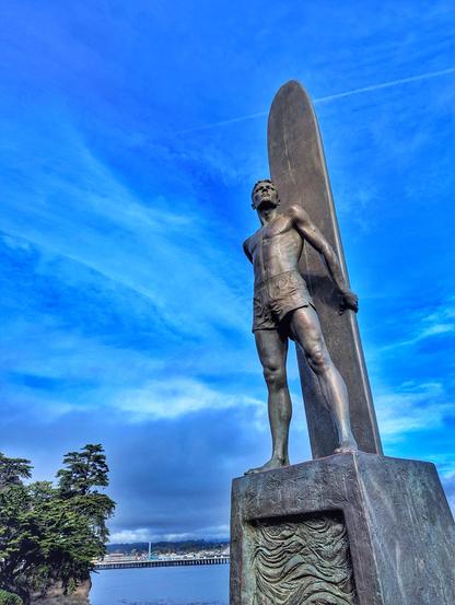 Looking upward at the surfer bro statue on a blue sky day.