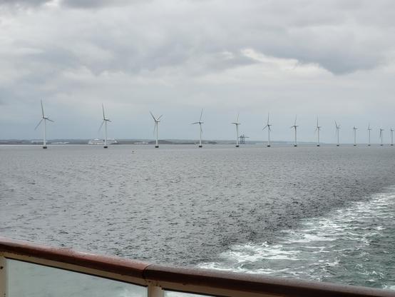 Rechts ist ein Teil der Heckwelle, und in der Mitte des Fotos stehen im Wasser mehrere Windräder. Hinter den Windrädern sieht man am Horizont noch einen Teil des Hafens von Kopenhagen. Der Himmel ist komplett bewölkt.