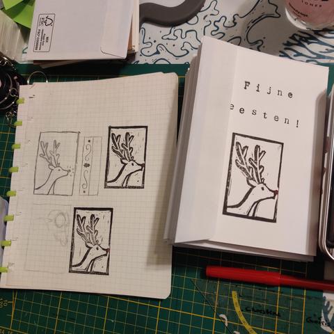 A cutting mat with some misc stuff on it (a protractor triangle, a red marker, some envelopes and keychains etc) plus a sketchbook with squares that contains sketches (of the reindeer among others) and test prints of the final thing, which is next to that: white holiday cards with envelopes under each card. The cards are printed with "Fijne feesten!" in black ink, with a linoprint of Rudolph underneath. He is printed in black, but his nose has been coloured red.