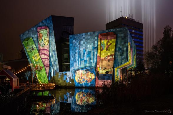 The Van Abbemuseum, with a colourful projection on it. Sort of a child’s drawing (but could also be a famous artist). Behind that a building, behind which light beams are projected to the sky. 