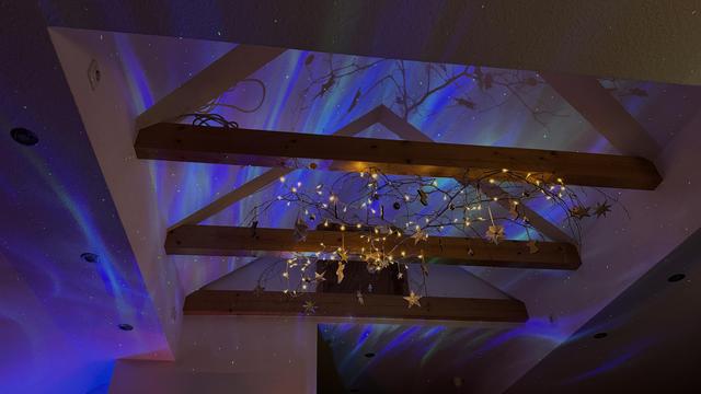 A starlight and Aurora Borealis projector as well as well as a string of lights hanging from a large branch atmospherically illuminate the open roof beams and pointed roof of an attic flat. 