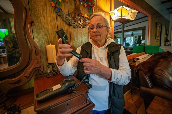 Older woman in a cozy living room holds a corded landline phone handset, inspecting it with concern as she stands beside a wooden table and lamp.