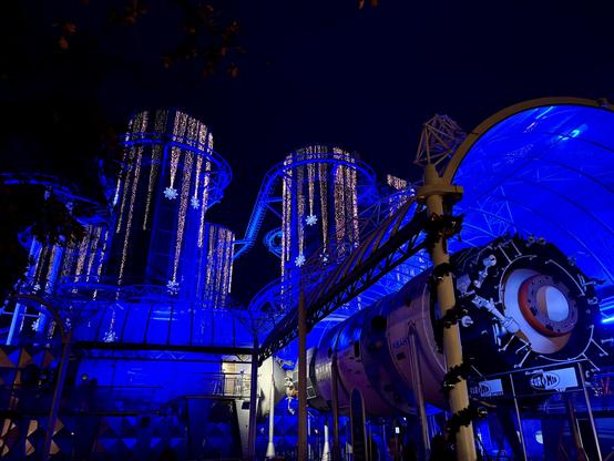 At night in the background, the EuroMir coaster lit up with Christmas lights; in the foreground, on the ground, a life‑size replica of the EuroMir space station glowing in blue/purple light.