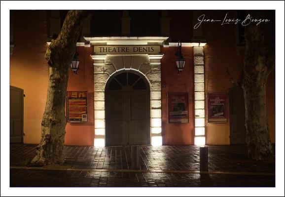 A small theater façade glows softly against the night. The building’s wall is painted a warm, earthy orange, which makes the illuminated stone doorway stand out like a stage within a stage. Four narrow columns of light rise from the ground, washing up the pale stone frame and emphasizing each block, as if the architecture itself is being spotlighted.
At the center is a tall, arched double door in deep shadow, closed and slightly mysterious. Above it, a simple sign reads “THEATRE DENIS,” held within a classical stone entablature that hints at old-world European design. Two traditional lantern-style lamps flank the doorway, hanging from ornate brackets; they’re unlit, leaving the main lighting to the dramatic footlights below.
Posters in vivid reds and yellows are mounted on both sides of the entrance, suggesting upcoming plays or events, adding splashes of color and a sense of life to the quiet street. The cobblestone or tiled pavement in front reflects the lights in faint streaks, giving the impression of recent rain or just polished stone.
Two broad tree trunks frame the scene on left and right, their rough bark catching a little of the glow. They act almost like natural curtains pulled back from the “stage” of the theater entrance—a subtle visual echo of performance and framing.
