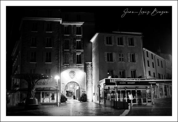 
The image is a black‑and‑white night photograph of a small European town square.

Key visual details:

Tall, old stone buildings dominate the scene, with many shuttered windows. Light from inside a few windows creates small bright rectangles against the dark facades.
At the center is a large arched passageway through one building, glowing with light from within and leading to a dimly visible street or courtyard beyond.
Street lamps cast pools of light on wet pavement, suggesting recent rain; reflections shimmer on the ground.
On the right is a corner café or bar with a striped awning and illuminated signs, adding a modern touch to the older architecture.
On the left is a sculptural tree-like form, perhaps a metal artwork or unusual street fixture, standing in partial shadow.
The strong contrast between darkness and the lit archway can symbolize transition, invitation, or passage from the unknown (dark square) to another, possibly safer or livelier space.
The empty square at night, combined with wet streets and historic buildings, evokes solitude and quiet reflection, a common theme in urban night photography.
Black and white emphasizes texture, light, and shadow over color, giving the scene a timeless, almost cinematic quality.