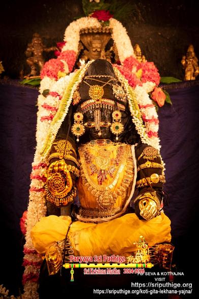 Closeup of idol of Hindu deity Krishna in yellow/amber seated in a relaxed yoga pose, right hand in blessing posture, left hand on left lap; wearing gold shoes, gold palm coverings, and lots of gold ornaments. A string of white flowers (some thin bands of red flowers) around Krishna. Dark blue (almost indigo) background. Other small metal idols on a ledge in the background.