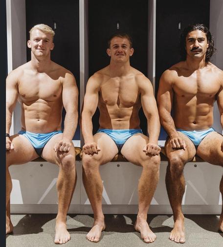 3 rugby players sit together on a bench in a locker room