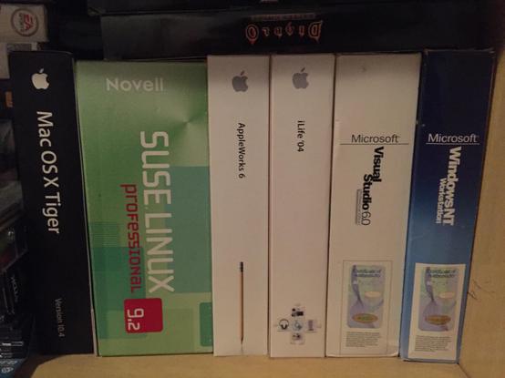 A shelf of boxed software. From left to right, there’s a black box of Mac OS X Tiger, a green box of SUSE Professional, a white box of Apple iWorks 6, a white box of iLife ‘04, a white box of Visual Studio 6, a blue box of Windows NT.