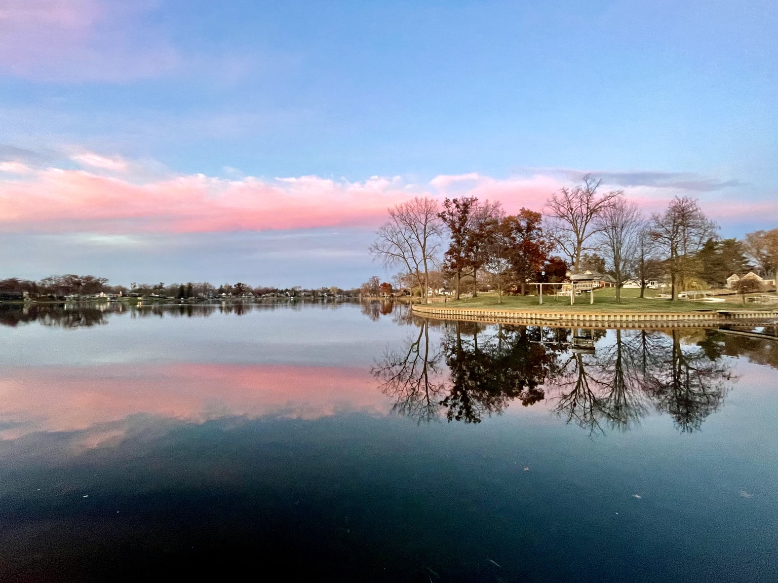 Sunset photograph of a low horizontal, pink cloud reflected in the lake. There is a small island to the right with some bare trees, and some trees with fall color leaves.