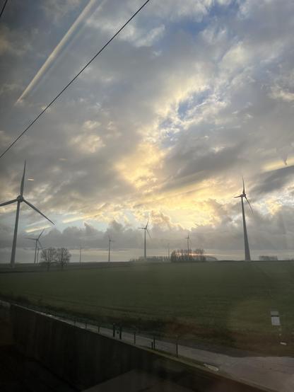 A scenic view from a train window featuring wind turbines in a grassy field under a dramatic sky with clouds and a sunset.