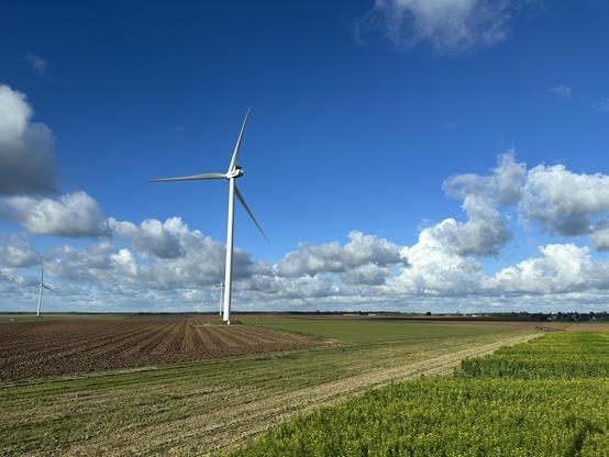 A landscape featuring a wind turbine amidst fields of varying crops. The sky is bright blue with scattered clouds. The foreground shows freshly plowed earth and green vegetation, while additional turbines are visible in the background.