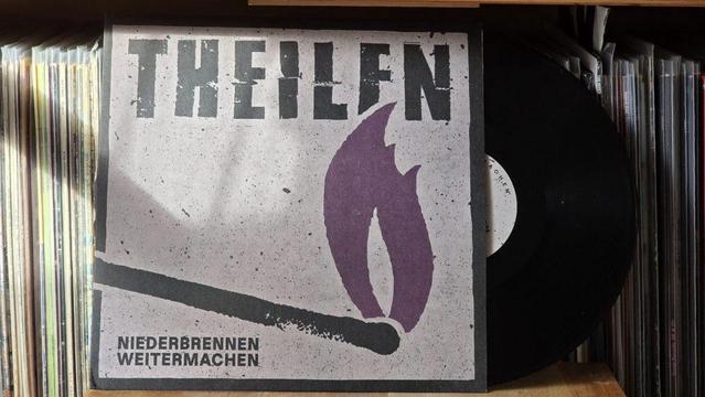 Ein Albumcover mit dem Titel „THEILFN“ und dem Untertitel „NIEDERBrennen WEITERMACHEN“. Das Design umfasst eine Streichholzschachtel und eine Flammengrafik vor einem strukturierten Hintergrund. Daneben liegt eine Schallplatte.
