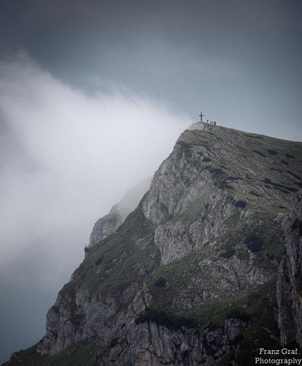 A dramatic mountain peak rises majestically against a moody, cloud-filled sky, its rugged cliffs partially shrouded in mist. At the summit, a solitary cross stands as a beacon, marking the highest point of the climb. A small group of hikers gathers near the cross, their figures silhouetted against the expansive view, emphasizing the sense of achievement and awe that comes with reaching such a breathtaking vantage point.

The steep, rocky slopes of the mountain are covered in patches of green gr…