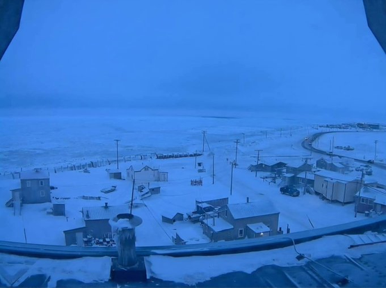 A wide-angle, cold-toned photograph of a small, snow-covered settlement in an arctic landscape. Scattered homes and utility poles are visible near a road that curves away towards the distant, flat, frozen sea or land under a solid blue sky.