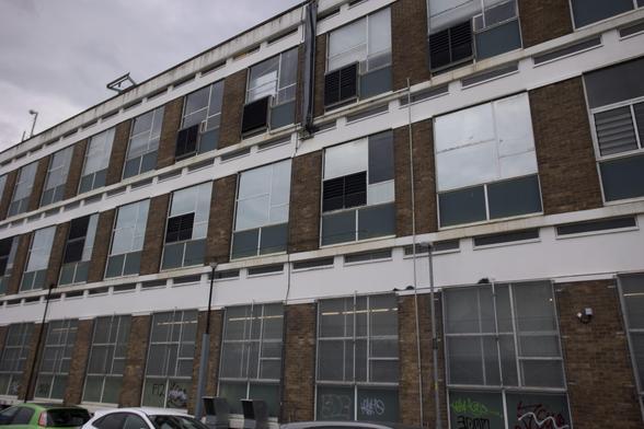 A three-storey building with brown brick and white trim, many large windows, some open or partially covered. The ground floor has graffiti on panels and several parked cars are visible in front. The sky is overcast.