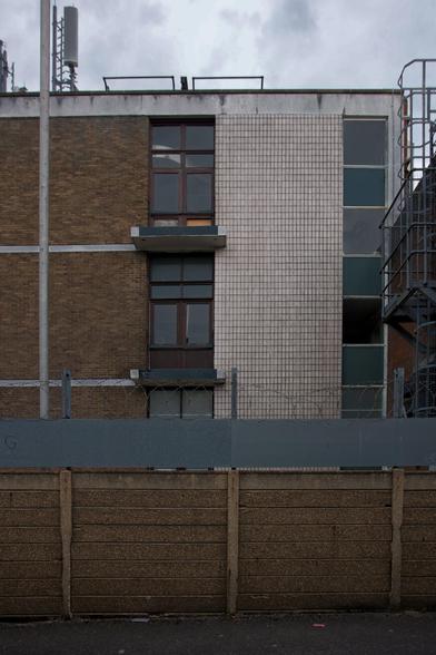 A three-storey building with brown brick and white tiled walls, tall windows, a metal staircase on the right, and aerials on the roof. The front is blocked by a high concrete and metal fence topped with barbed wire under a cloudy sky.