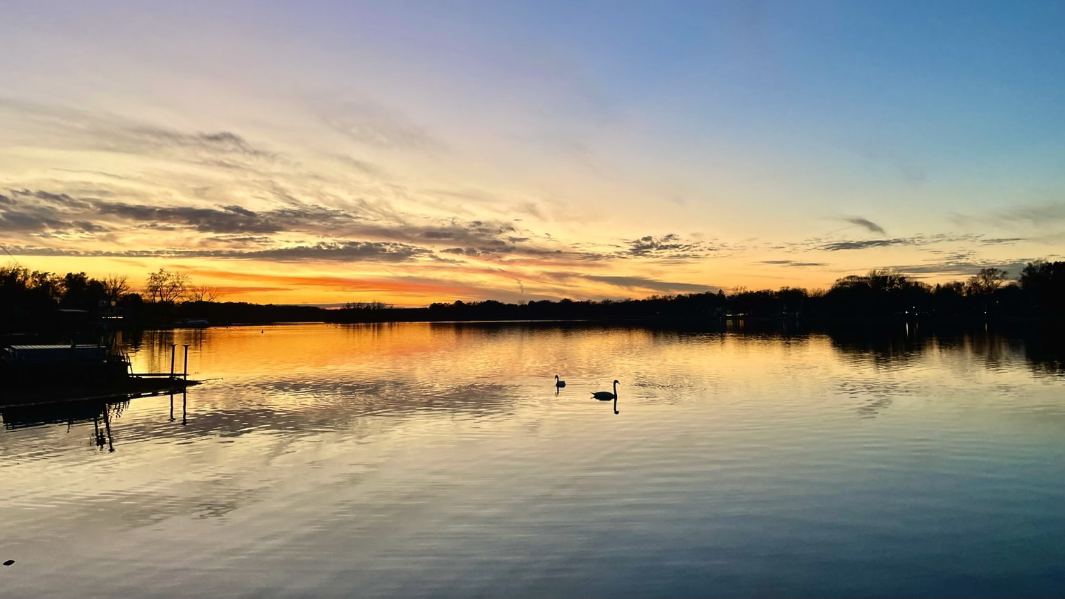 Sunset photo of a sunset behind a smooth reflective lake with orange light on the horizon and a pair of swans in silhouette in the middle distance.