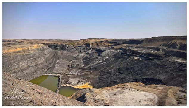 A panoramic, wide angle photograph in colour, signed by Kev Peirce, showing a massive open cut mine.
The image captures the immense scale of the excavation, with distinct, stepped layers (benches) visible along the sides of the pit, highlighting the geology and mining method.
The base of the mine is a mix of dark, excavated material and two pools of stagnant, yellowish green water. The surrounding landscape is arid and flat under a clear, pale blue sky, emphasising the isolation and the environmental impact of the operation.