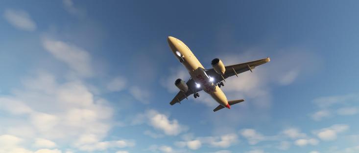 The image depicts a commercial airplane in flight against a backdrop of a clear blue sky with scattered white clouds. The aircraft is captured from a low angle, looking up at it as it ascends or cruises at a high altitude. The airplane's landing gear appears to be retracted, indicating it is in flight. The aircraft has a sleek design with two engines mounted under the wings, and the tail features a logo or text that is partially visible. The overall scene conveys a sense of travel and aviation.