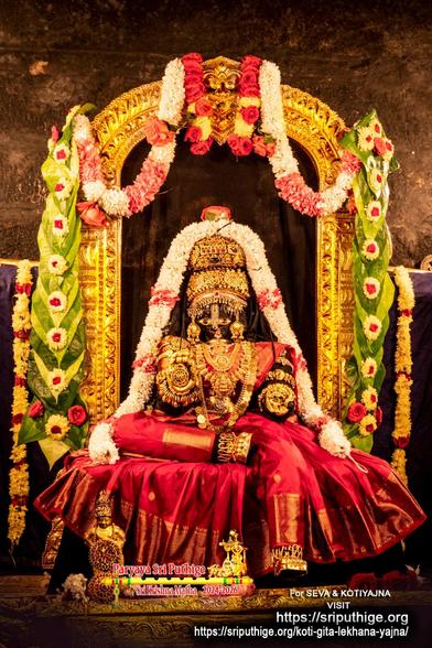 Idol of Hindu deity Krishna dressed in red as Lakshmee, left leg down, right leg folded flat and placed on the seat, right sole barely touching the left inner thigh; wearing gold shoes, gold palm coverings, a 2-level gold crown, and lots of gold ornaments. A string of white flowers around Krishna, the red saree covering the seat. A gold arch behind Krishna. A string of green betel leaves (yellow and red flowers on top of each leaf) hangs down each side of the arch. At string of red and white flowers hooks for the left top of the arch to the middle. Likewise on the right. Earthen red-brown background wall.
