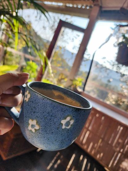 Photo shows a speckled blue ceramic cup with white daisies on it, being held up by a hand.

There is steaming hot coffee in the cup.

A patio framing greenery and a mountain range are visible, out of focus, beyond the cup.