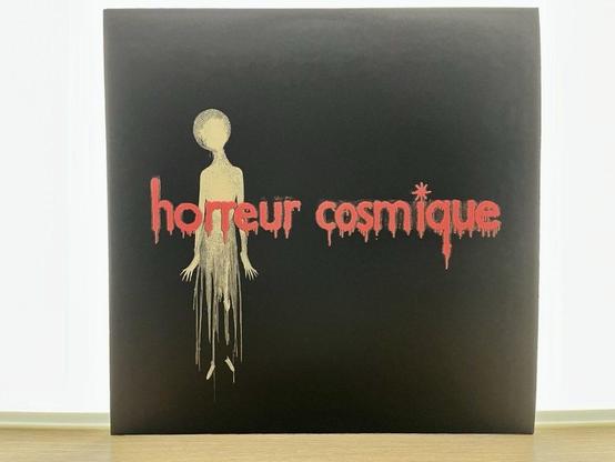 Ein schwarzes Albumcover mit dem Schriftzug „horreur cosmique” in roten, tropfenden Buchstaben. Eine abstrakte Figur mit rundem Kopf und langgestrecktem Körper ist in Gold dargestellt und befindet sich in der Mitte.