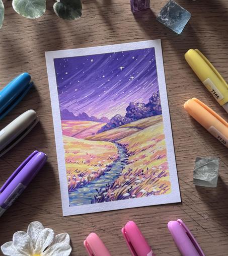 A photo of a portrait-format painting, placed on a wooden desk and surrounded by acrylic markers, fluorite crystals, flowers, and leaves. The painting features a roughly paved pathway leading through yellow, grassy fields with pink/purple shadows, under a purple starry sky.