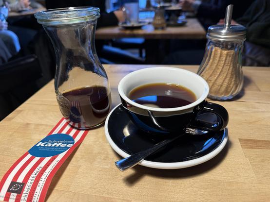 Eine Kaffeetasse auf einem Holztisch neben einer Karaffe gefüllt mit leckerem Filterkaffee. Ein Zuckerstreuer ist ebenfalls im Bild, wird aber nicht gebraucht.