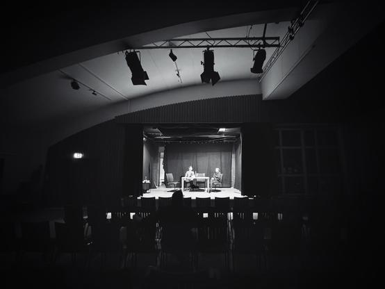 S/W-Aufnahme eines Theatersaals mit einer beleuchteten Guckkastenbühne im Hintergrund. Der Rest des Raumes ist dunkel.