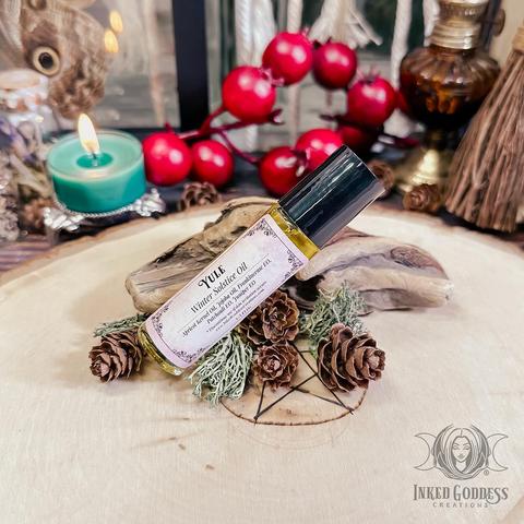 A small round vial of yellowish "Yule / Winter Solstice Perfume Oil" is propped on a raw wood slice. It has a black cap and a label that lists its ingredients, which include apricot kernel oil, jojoba oil, and frankincense, patchouli, and juniper essential oils. This is an exclusive product from Inked Goddess Creations.