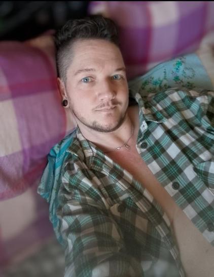 Trans man with blue eyes and brown hair and facial hair. He wears a plaid shirt in green and white, and reclines on pink and white plaid pillows. His shirt is open down the front, showing part of his chest. He has a slight smile and looks at you with intensity that borders on lustful.