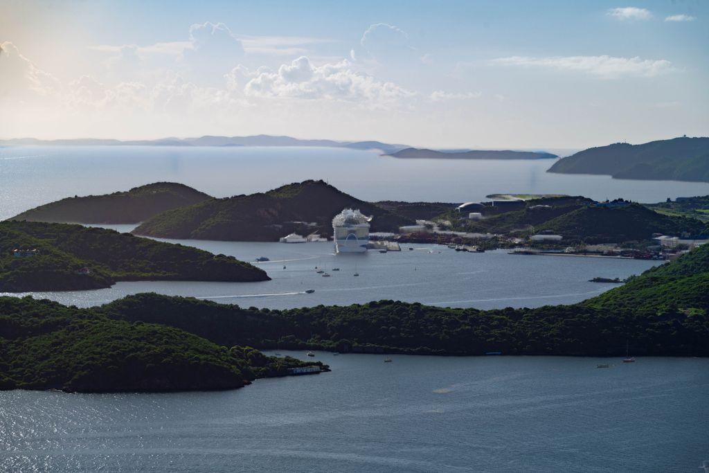 A high vantage view overlooking Charlotte Amalie harbor in St. Thomas, USVI. Green islands and peninsulas curve around calm blue water, with the large cruise ship Icon of the Seas docked in the distance amid scattered sailboats.