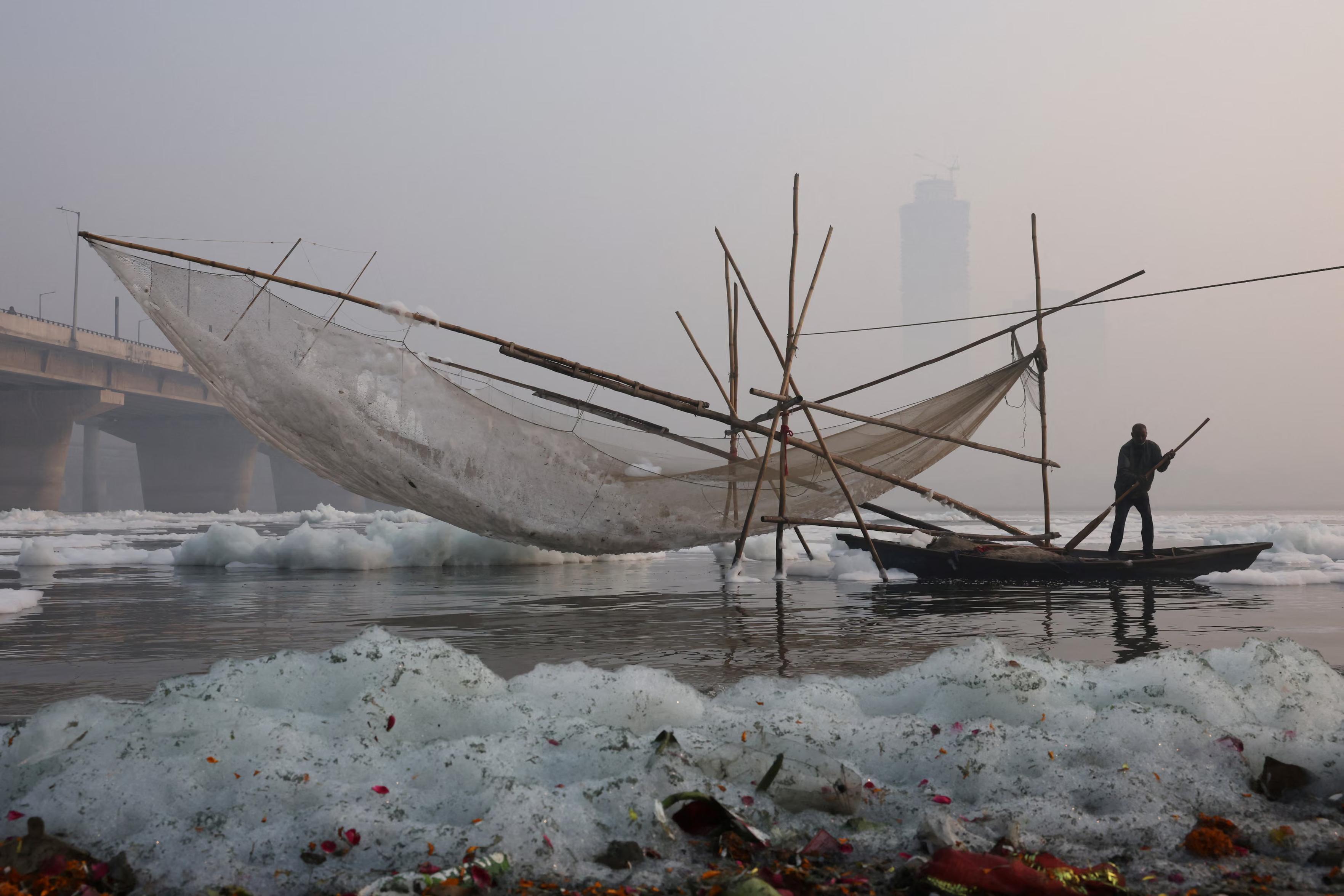 A fisher stands next to a net covered with foam in the polluted #Yamuna River on a smoggy morning.