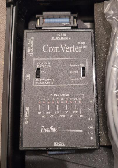 A Frontline "Comverter", a small plastic box, slightly bigger than the palm of a hand, with a flat ribbon cable at the bottom and a DB25 port on the top.