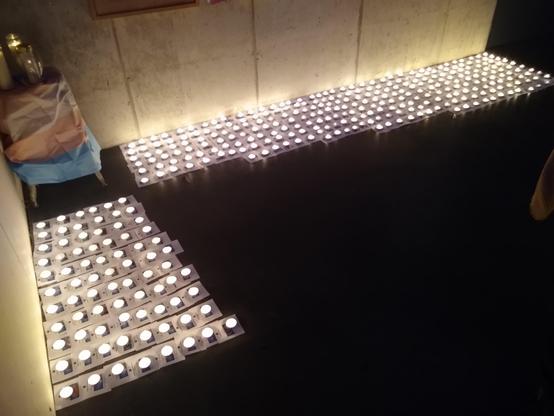 Photo d'un mémorial constitué des identités et photos des personnes trans assassiné•e•s ou poussé•e•s au suicide. Sur chaque vignette est déposée une bougie. Dans le coin, un drapeau trans entoure une console où sont posée trois grandes bougies