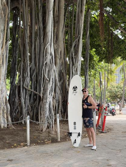 Me with a surfboard I didn’t ride because it’s too short at Waikiki beach in front of a massive tree