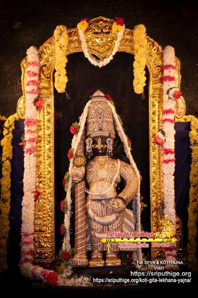 Idol of Hindu deity Krishna standing in diamond cladding with a matching crown and a similarly clad staff in right hand. A string of white flowers (with intervening red roses) around Krishna. A gold arch around Krishna. A string of white flowers (some red flowers) hangs down along each side of the arch. A short string of yellow flowers hangs from the left top of the arch, and from the right top of the arch. Dark cloth filling between the sides of the arch. Dark cloth background behind the arch. A string of yellow flowers hangs down at each end of the background.