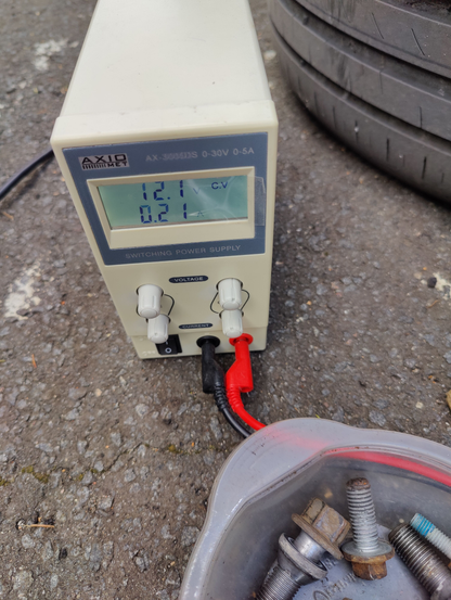 Bench power supply configure to 12V next to tire and bunch of bolts on a tarmac.