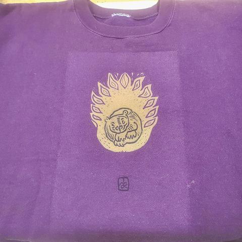 Un sweat violet, impression d'une grande flamme jaune, dans laquelle un petit ours, enroulé sur lui dans une position bizarre, regarde le public