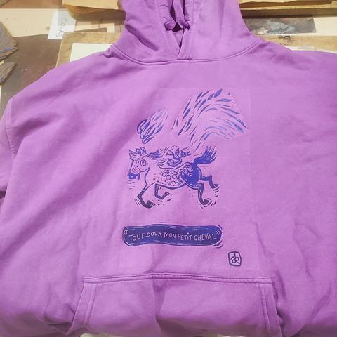 Un sweat à capuche violet clair, impression en mélange noir et bleu : un cheval galope avec un air hagard, sur son dos un petit personnage à bonnet d'âne valdingue vénère, d'ailleurs il dit "tout doux mon petit cheval". Mais bon il y a une flamme au-dessus de la tête du cheval et tout autour, à mon avis il ne va pas se calmer.